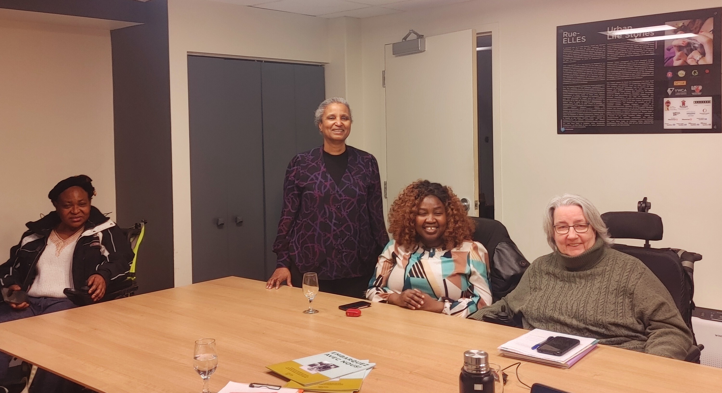 Nous voyons sur la photo 4 femmes qui ont pris part à la rencontre. Il y a Eveline Claire, Florence et Célia qui sont 3 femmes afro descendantes. Eveline est assise dans son fauteuil roulant, Florence est debout et Célia assiste. Manon est à droite souriante avec, une peau blanche, ses cheveux gris et son grand sourire. Elle est assise dans son fauteuil roulant. 